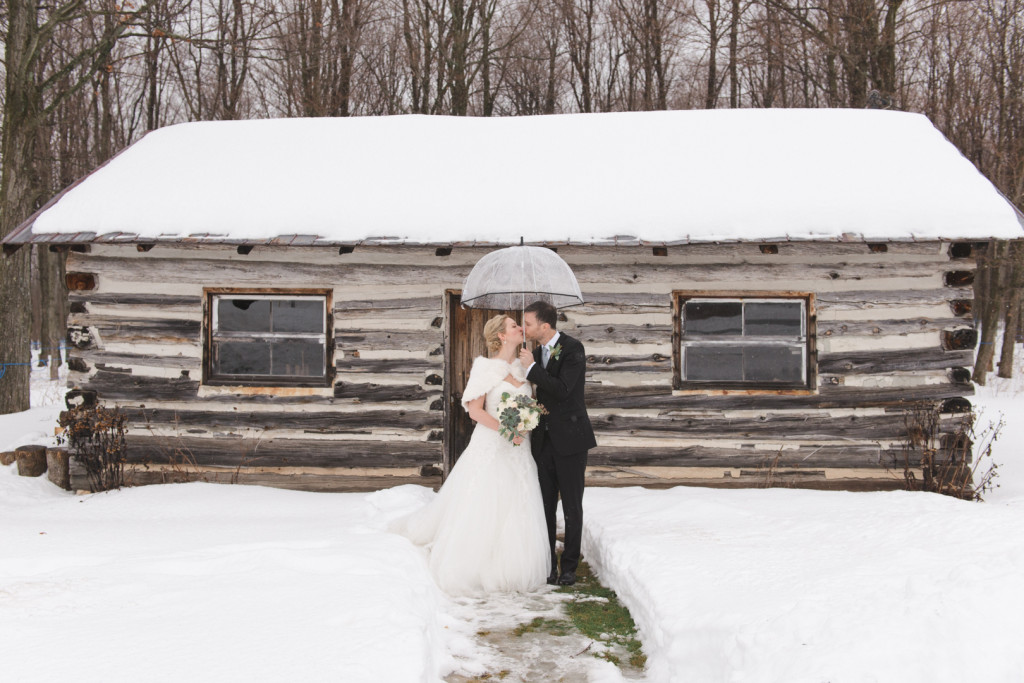 Bride and groom with umbrella at winter wedding in front of rustic shed