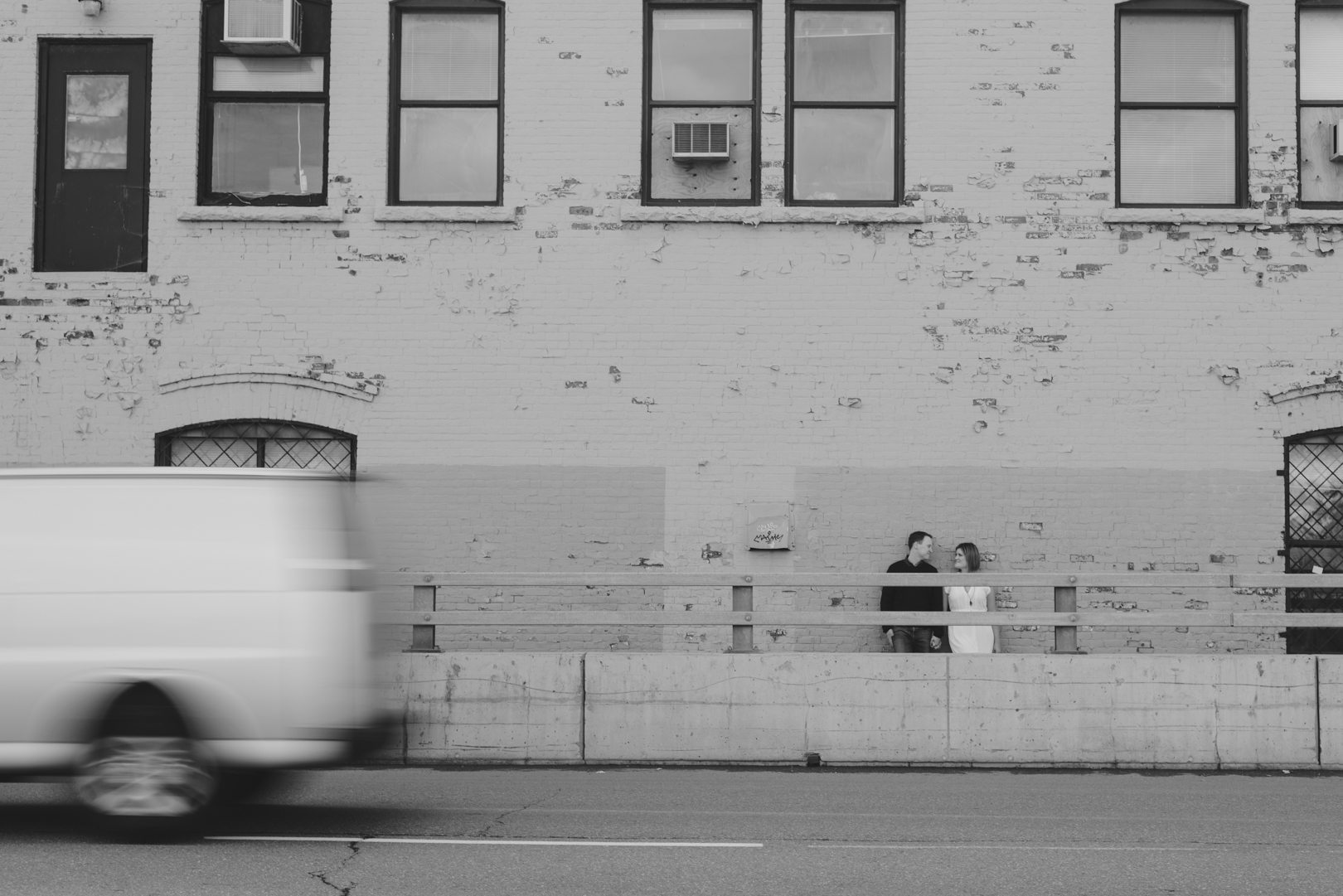couple standing against old brick building with white van driving past
