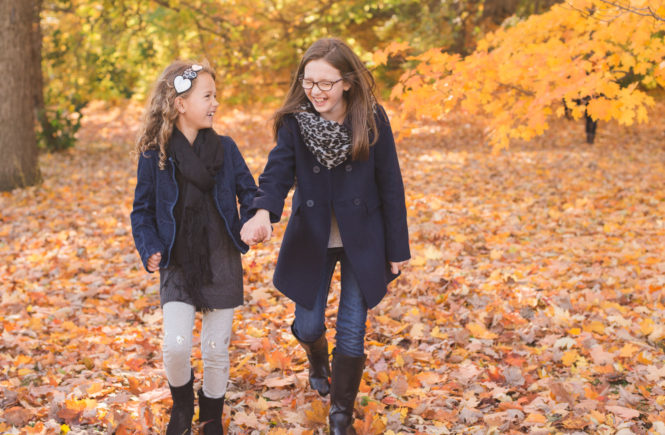 Two sisters holding hands walking in the fall leaves on a sunny day