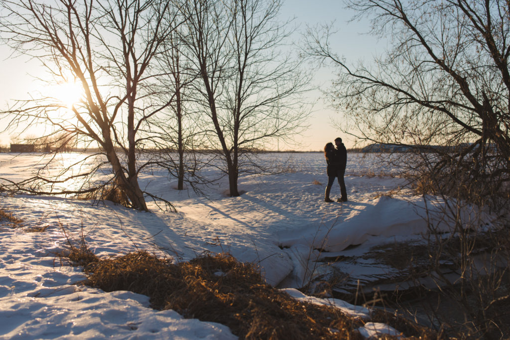 couple standing along snowy ravine among the trees at sunset