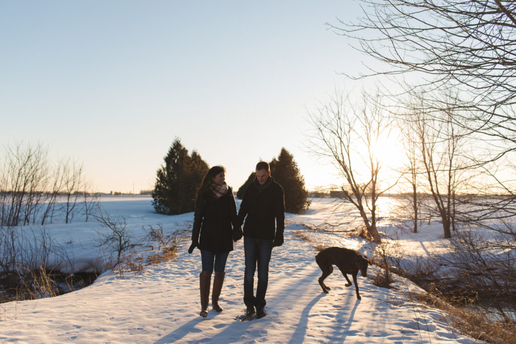 couple and their dog walking along snowy path at sunset