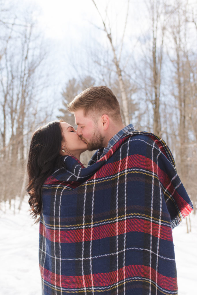 engaged couple cuddling up in a plaid blanket in winter