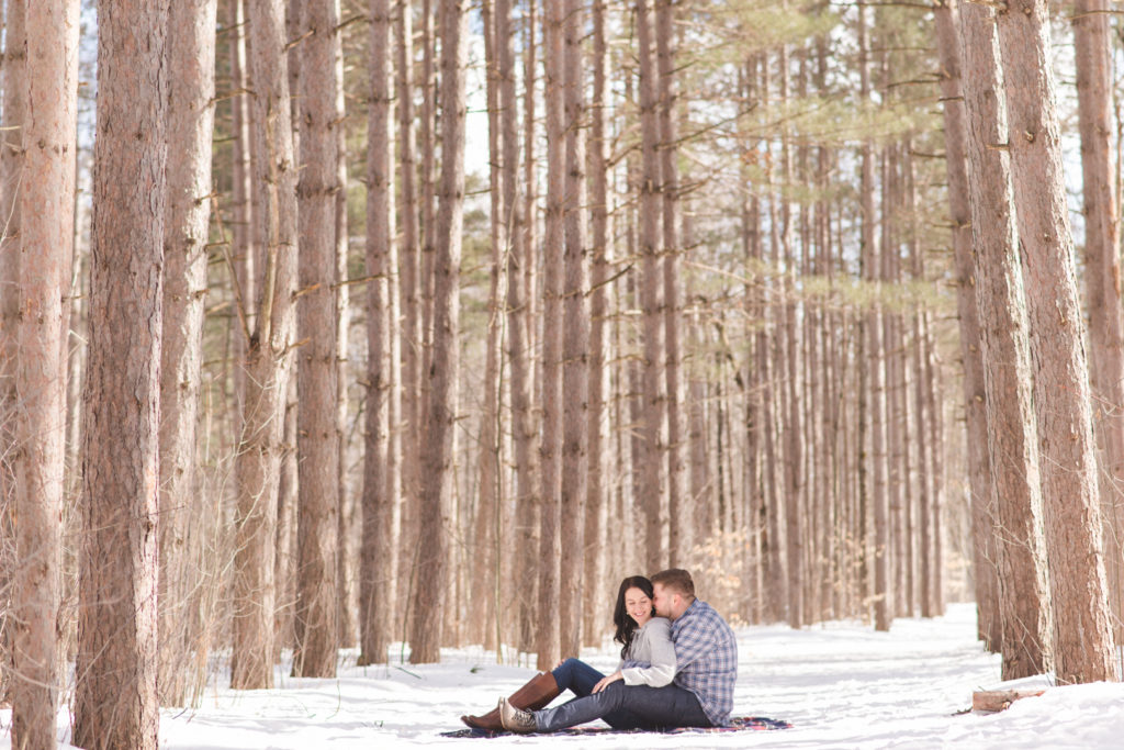 Engaged couple cuddled up on a blanket in the winter among the trees