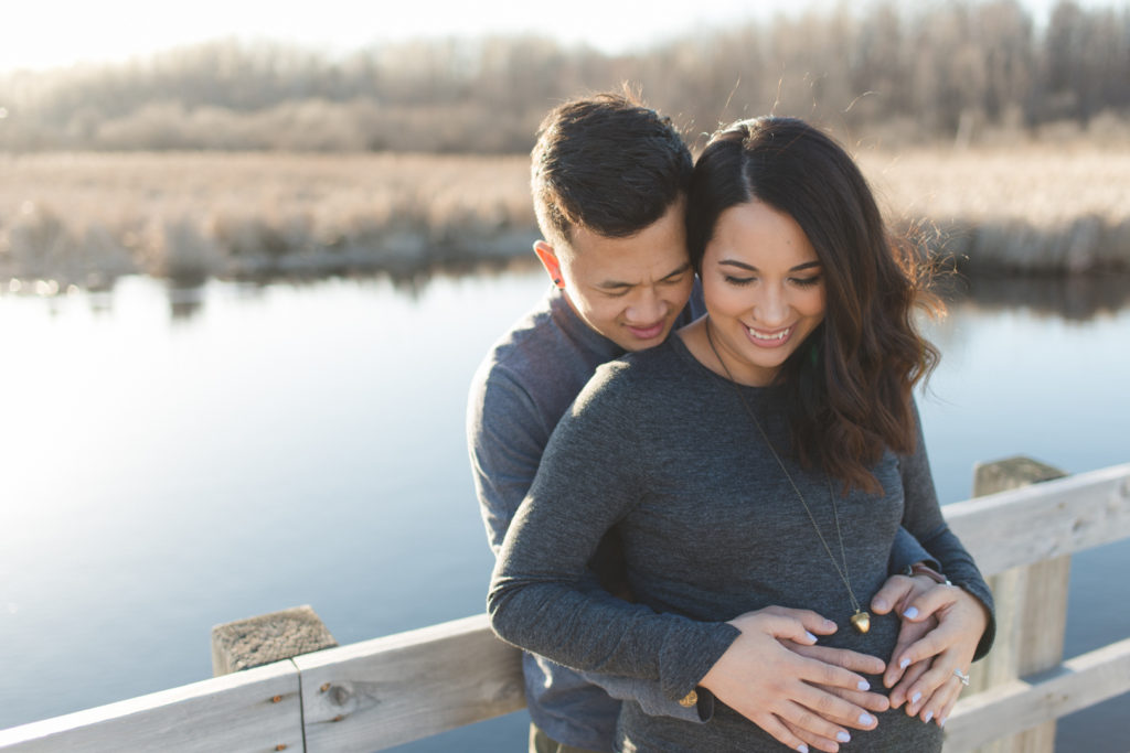 expecting parents standing along wooden bridge at sunset