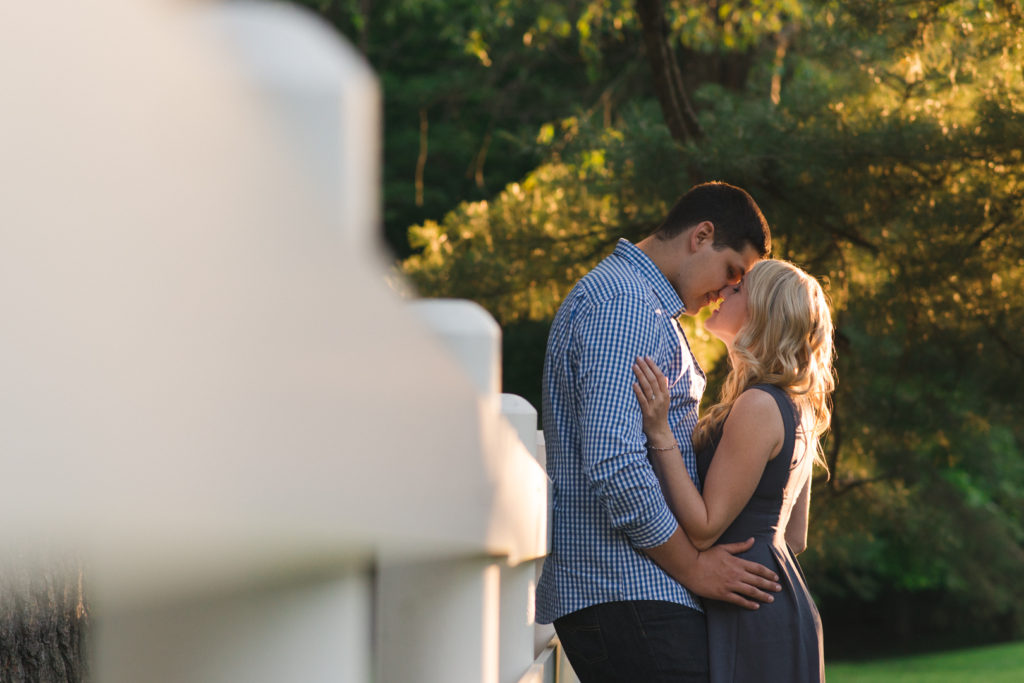 photo of engaged couple leaning up against a white fence cuddling at sunset