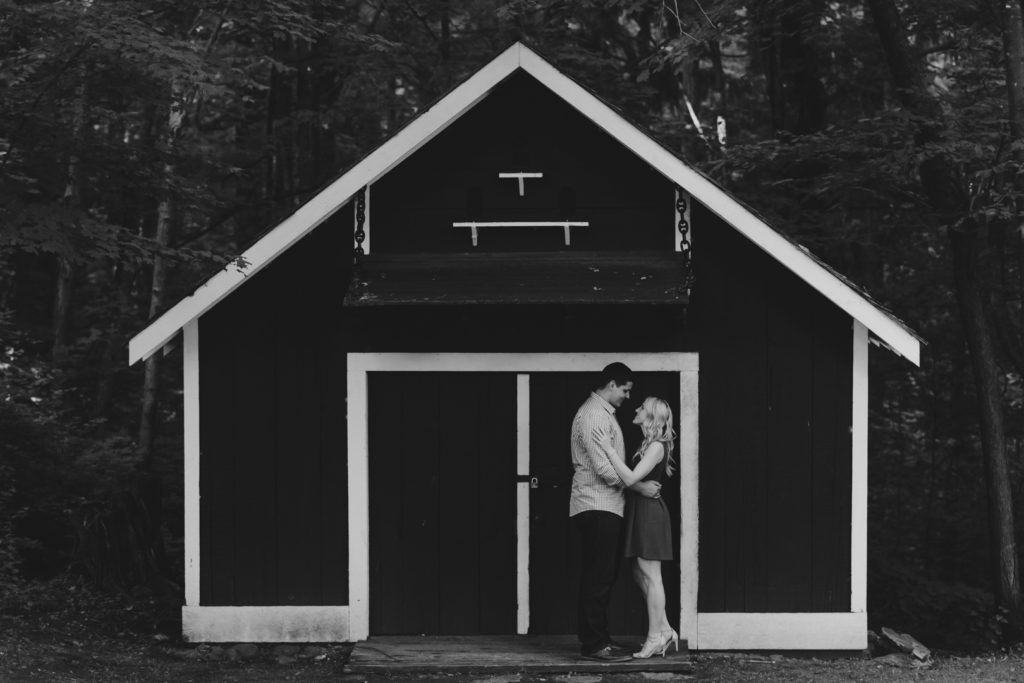 black and white photo of engaged couple cuddling in front of black and white shed