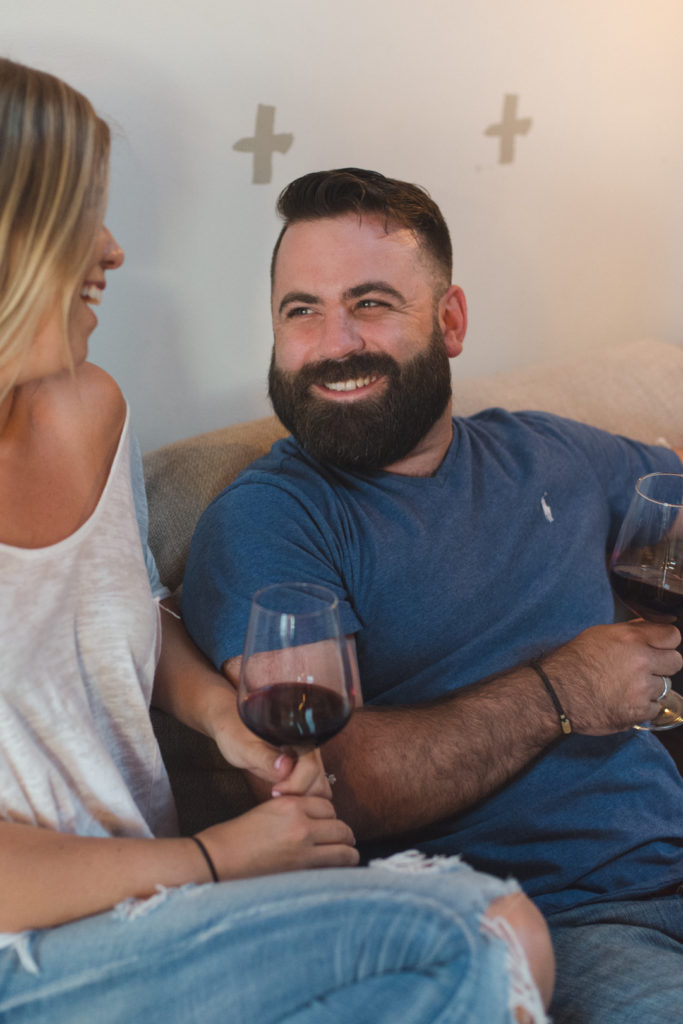 groom to be smiling at his fiancee as they enjoy a glass of wine on the couch