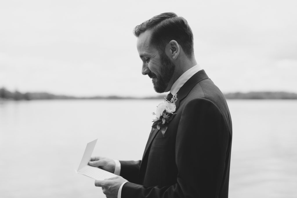 groom smiling as he reads a card from his bride on their wedding day by the water