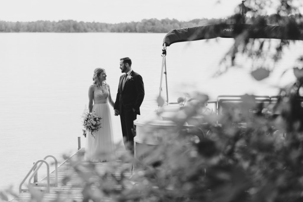 black and white photo of bride and groom standing on the dock by the lake