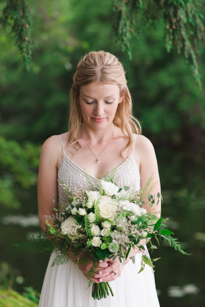 bride holding green and white bouquet standing under pine tree by the water