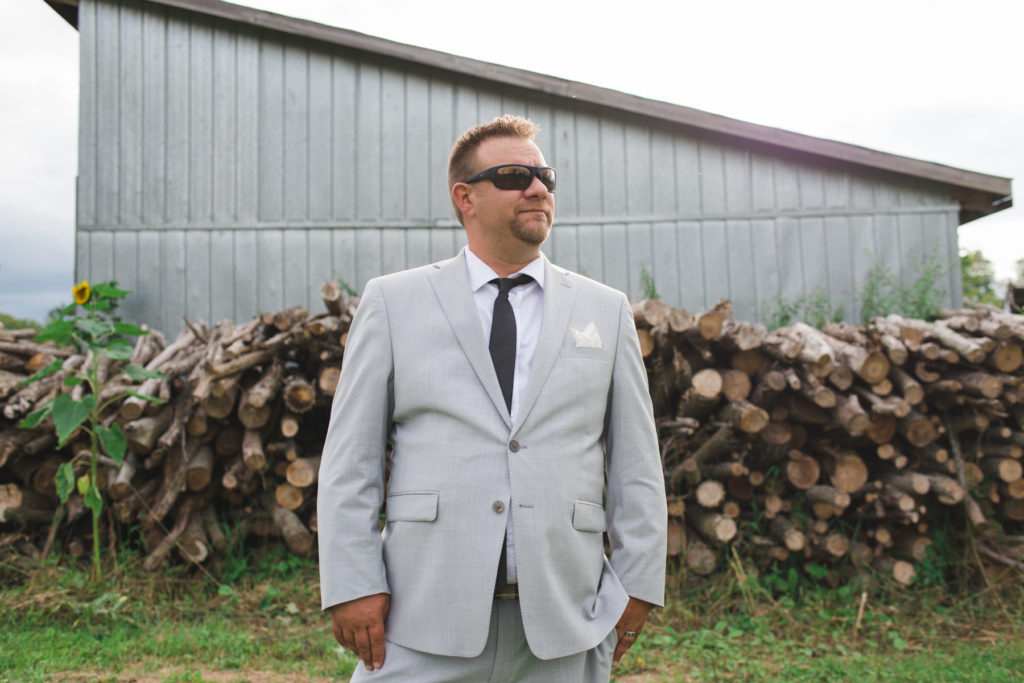 groom wearing sunglasses in front of pile of wooden logs