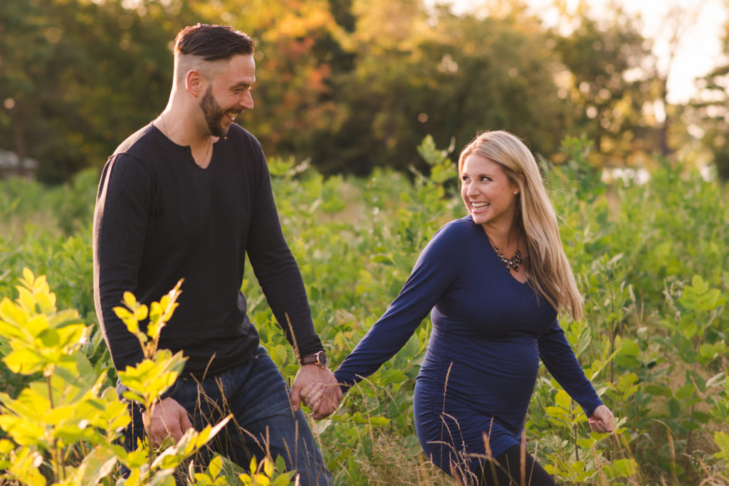 engaged couple expecting a baby walking in a field at sunset holding hands