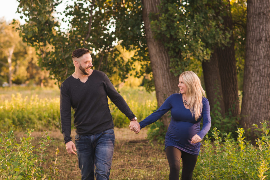 engaged couple expecting a baby holding hands as they walk through a field at sunset