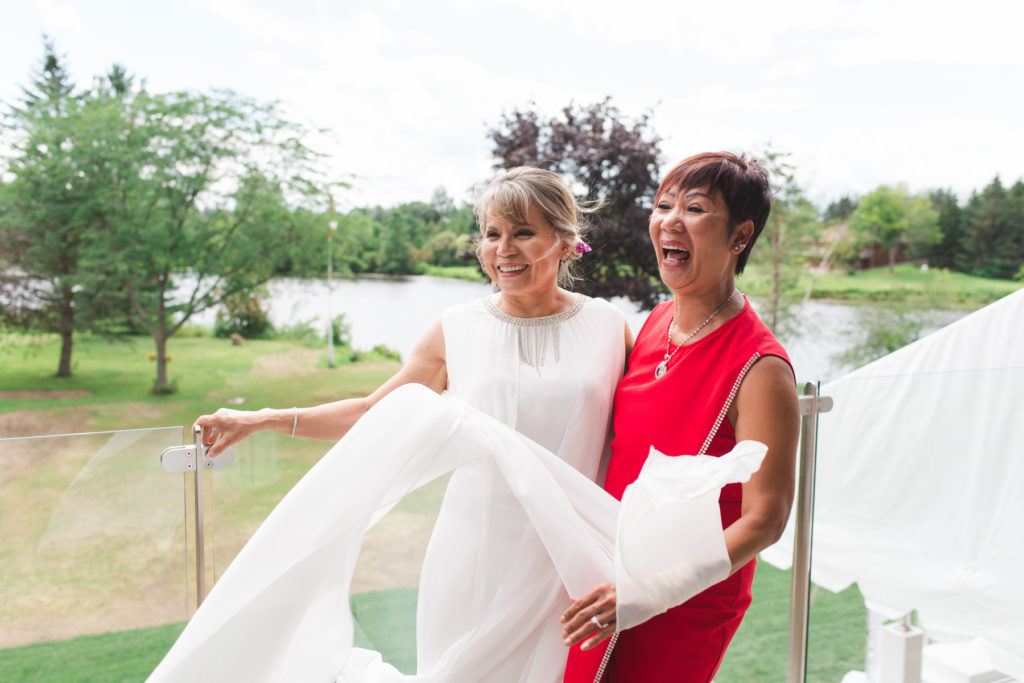 bride and friend laughing on balcony while dress blows in the wind