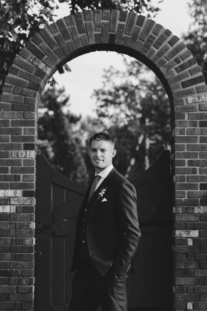 groom standing in front of stone archway in black and white