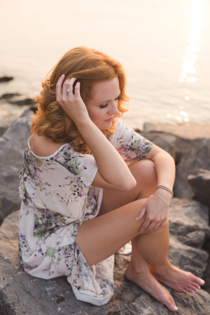 redhead girl sitting on rocks by the water at sunset