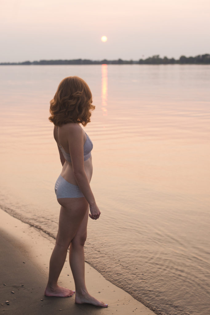 redhead girl standing by the water's edge in her undies at sunset