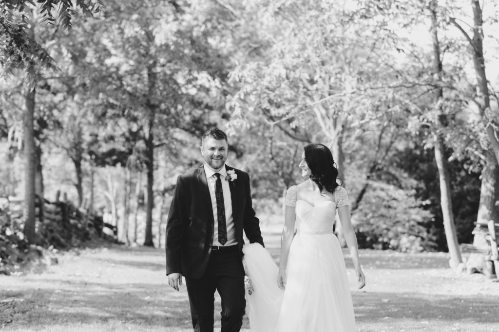 groom holding bride's train as they walk together along tree lined path