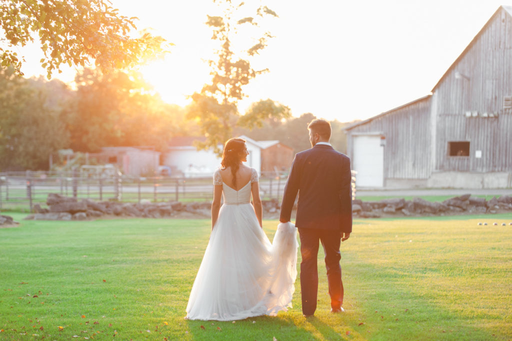 groom holding bride's train as they walk into the sunset on a farm
