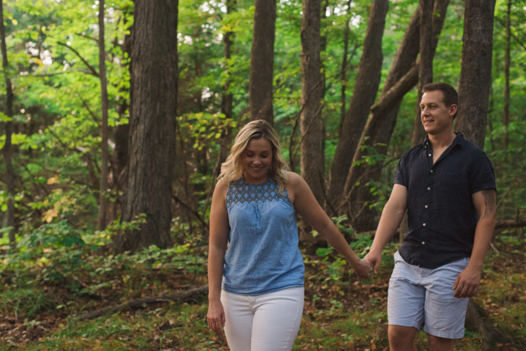 engaged couple walking along wooded trail holding hands