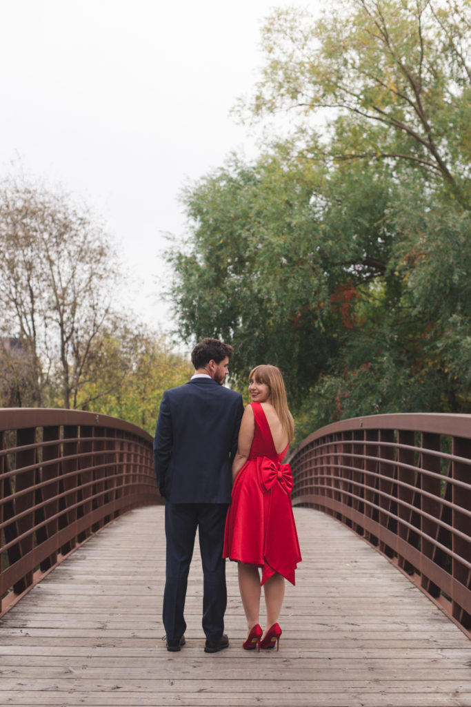 engaged girl wearing a red dress with a big bow standing on a bridge with her fiancee