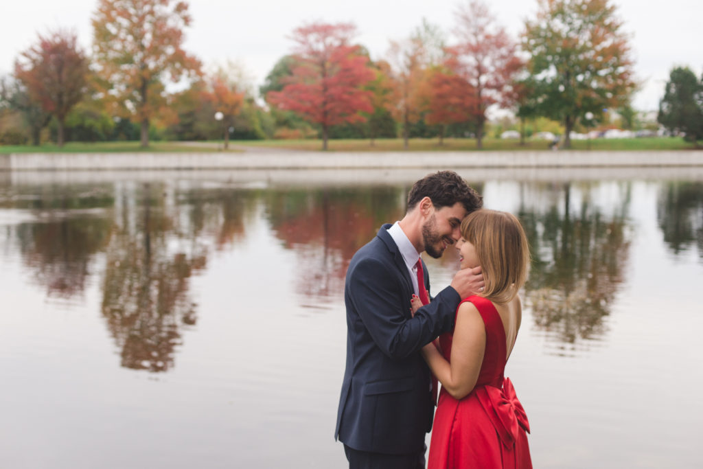 engaged couple kissing by the water with fall trees in the background