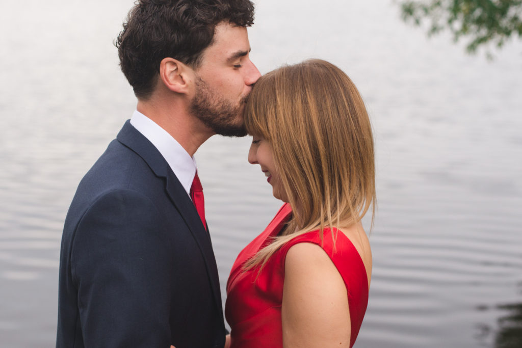 engaged man kissing his fiancee on the forehead by the water's edge