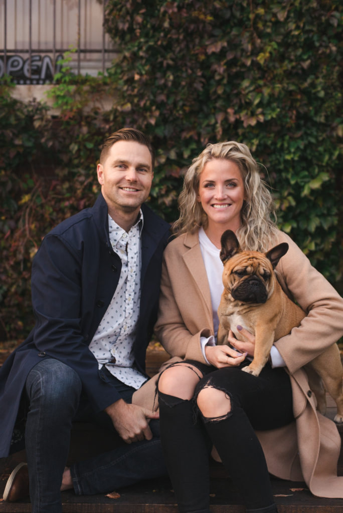 couple and their dog seated in front of vines with their dog