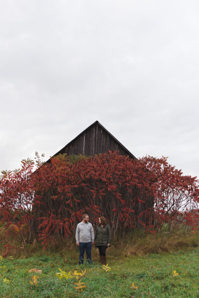 engaged couple standing in front of old barn with red trees