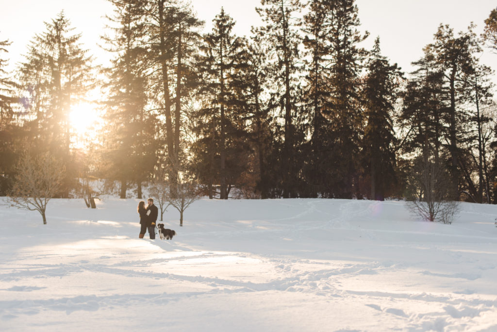 engaged couple standing in an open snowy field with their dog at sunset