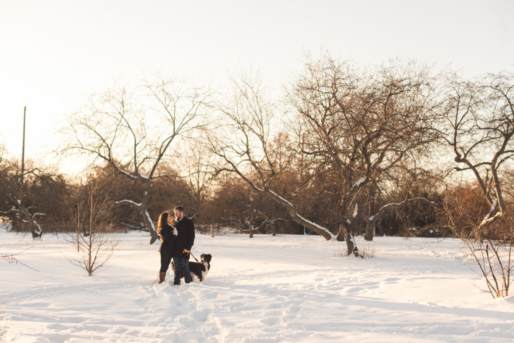 couple standing in a snowy field at sunset with their dog