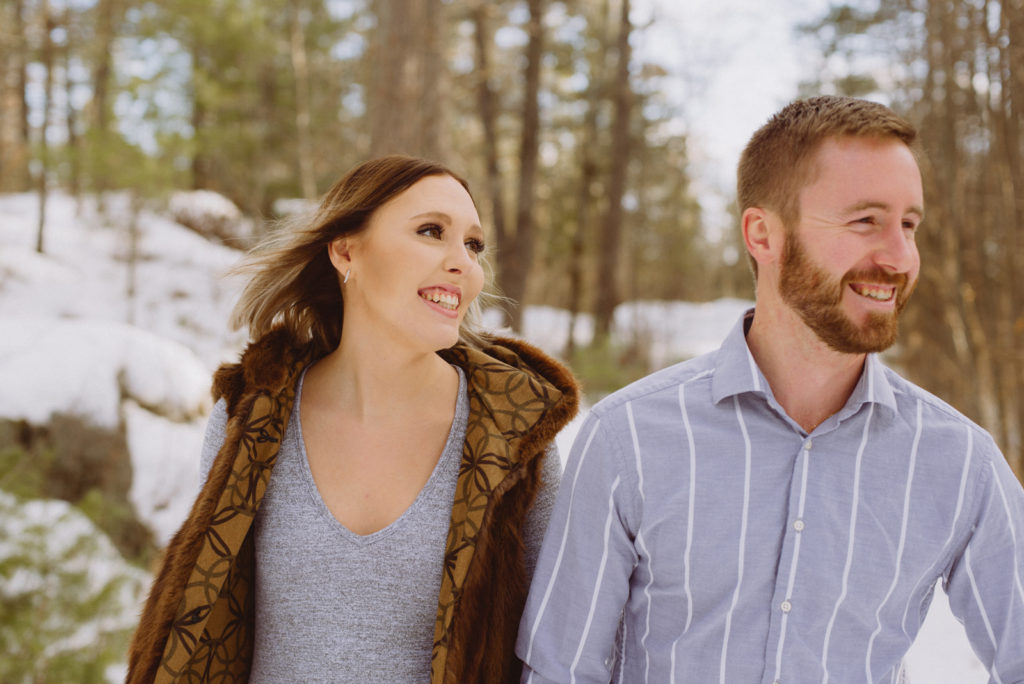 engaged couple laughing as they walk through the woods during winter