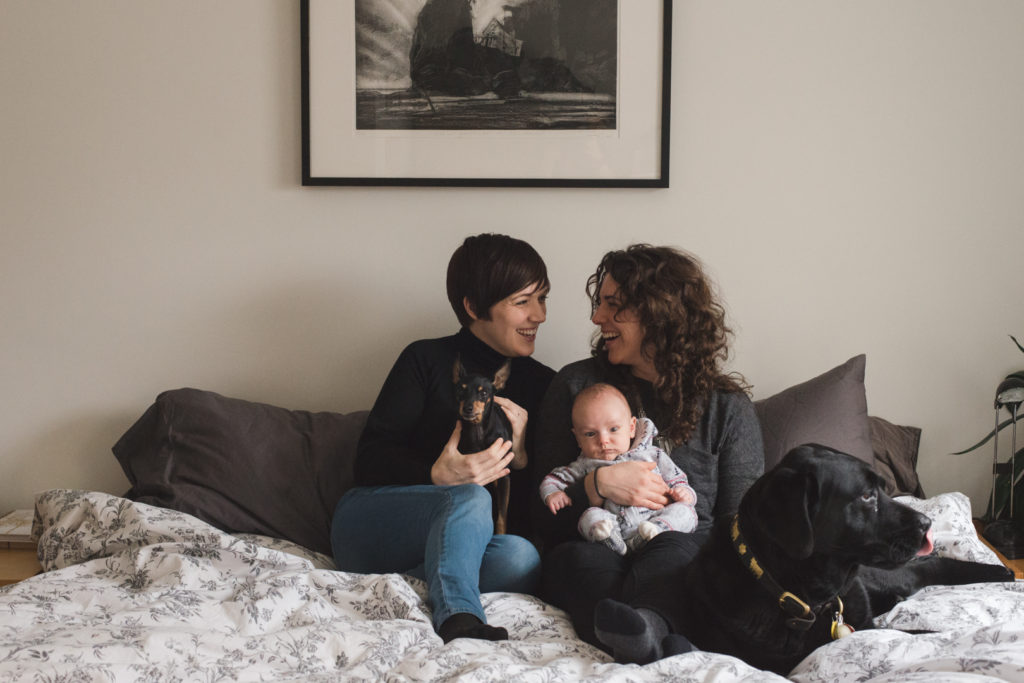 two moms laughing with each other with baby boy and dogs on their bed