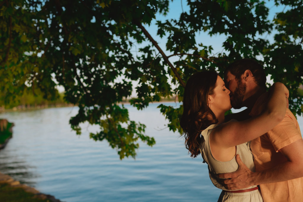 engaged couple kissing underneath a green tree by the water at sunset