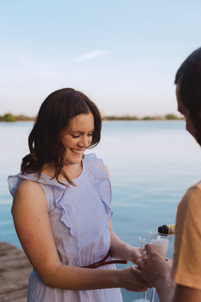 man pouring his fiancee a glass of champagne by the water at sunset