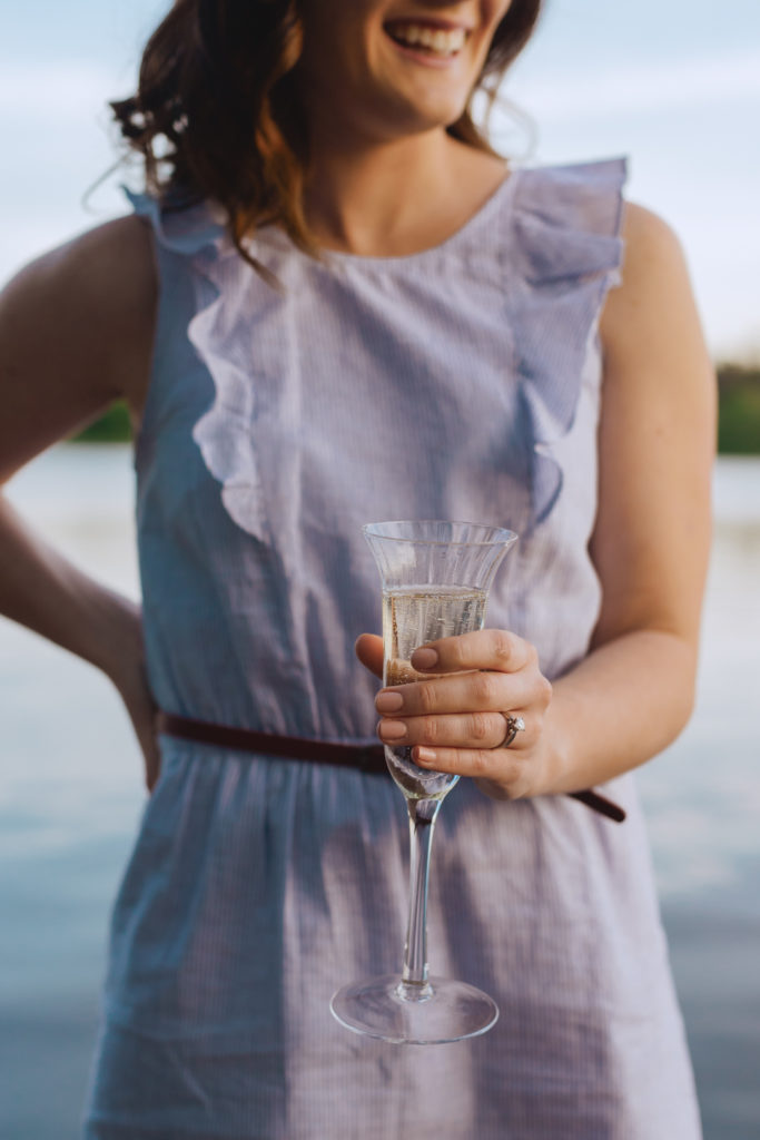 engaged girl holding a crystal champagne glass by the water at sunset