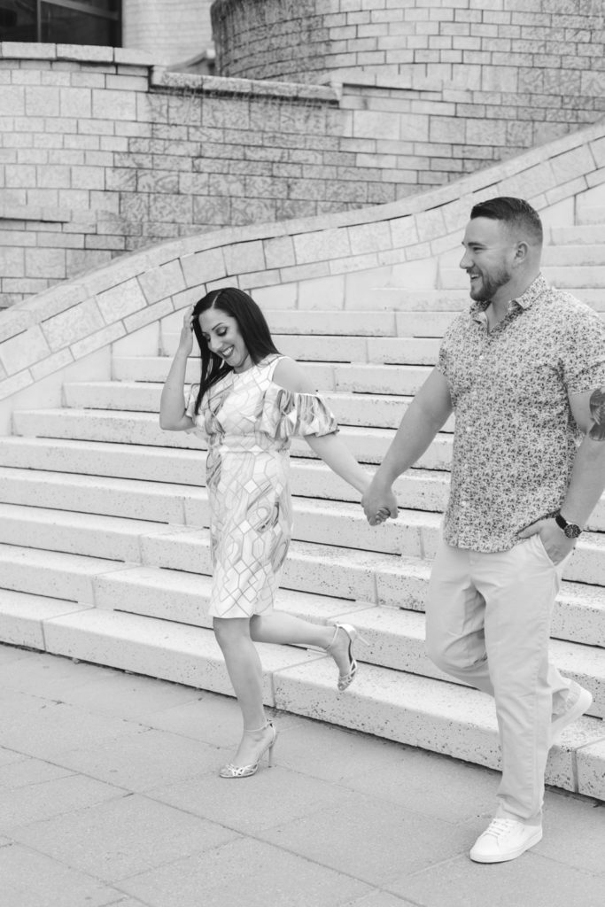 engaged couple walking down the steps holding hands and laughing in black and white