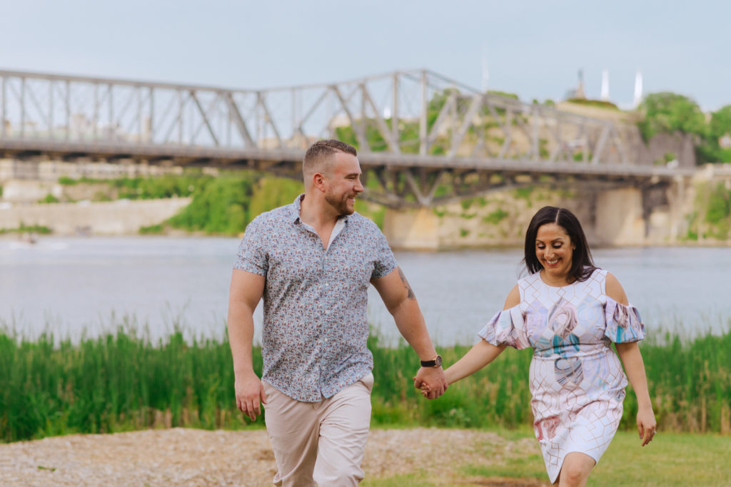 engaged couple holding hands walking by the Ottawa River and Alexandria Bridge