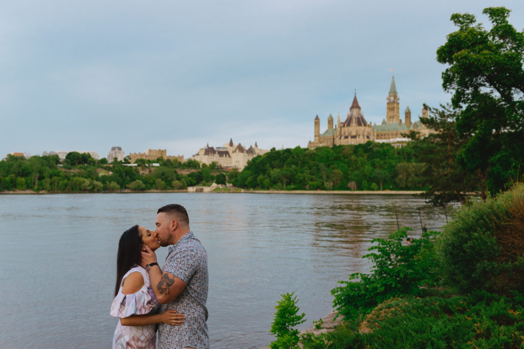 engaged couple kissing by the Ottawa River with the Parliament Buildings in the background