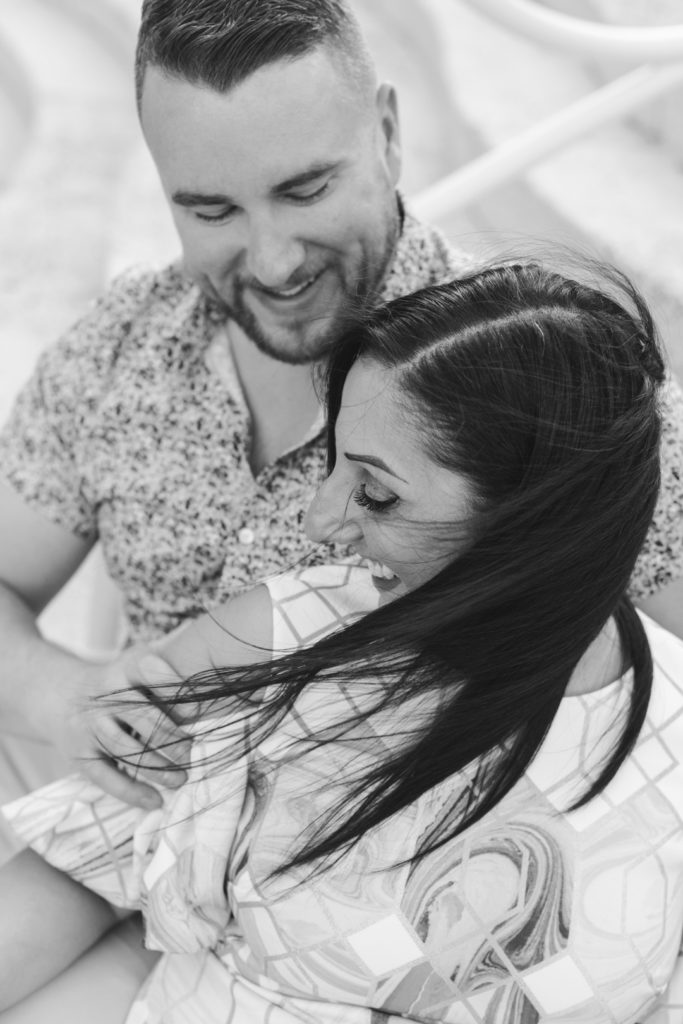 Engaged couple laughing sitting on the steps as the wind blows the woman's hair