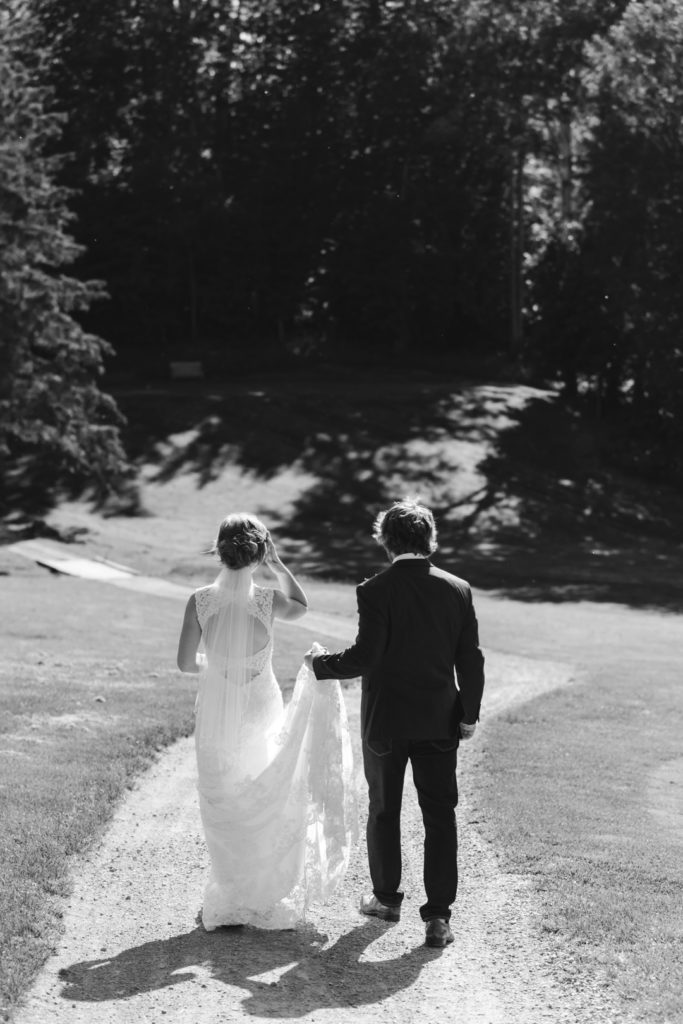 Groom holding bride's train as they walk down dirt path of golf course