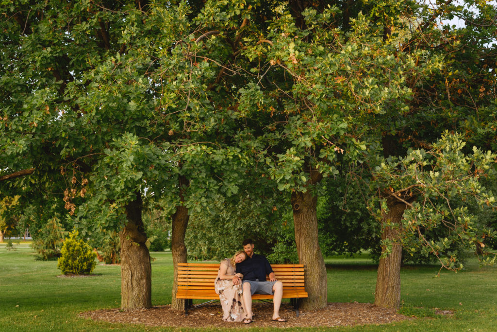 engaged couple sitting on a bench together underneath a tree at sunset