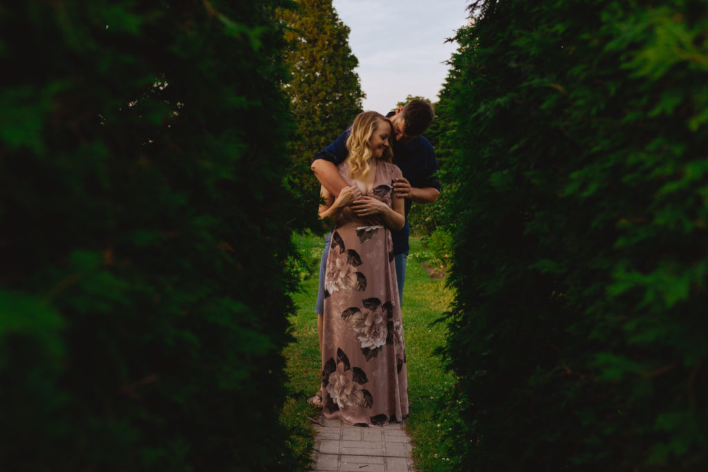 engaged couple with arms wrapped around each other between two cedar hedges at sunset