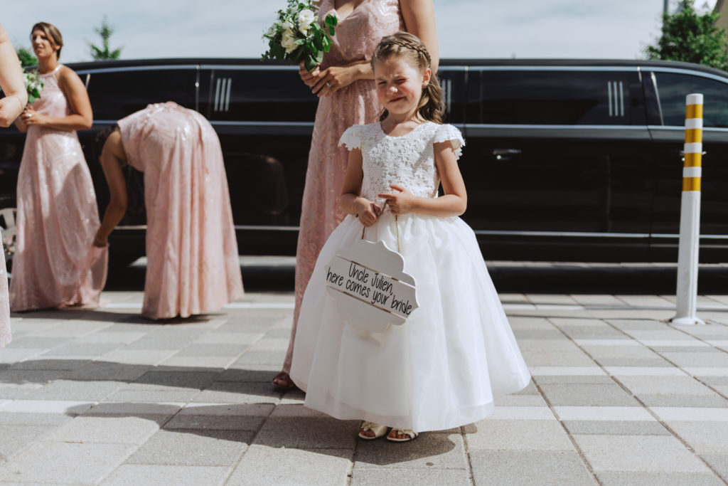 flower girl holding white sign that says 'here comes your bride' in front of black limo