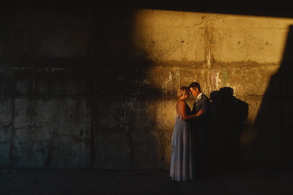 bride and groom standing in patch of orange sunlight at dusk against brick wall
