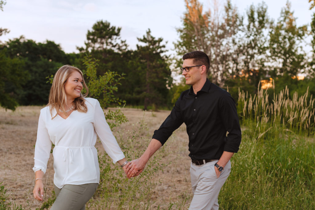 engaged couple walking in a field at sunset holding hands