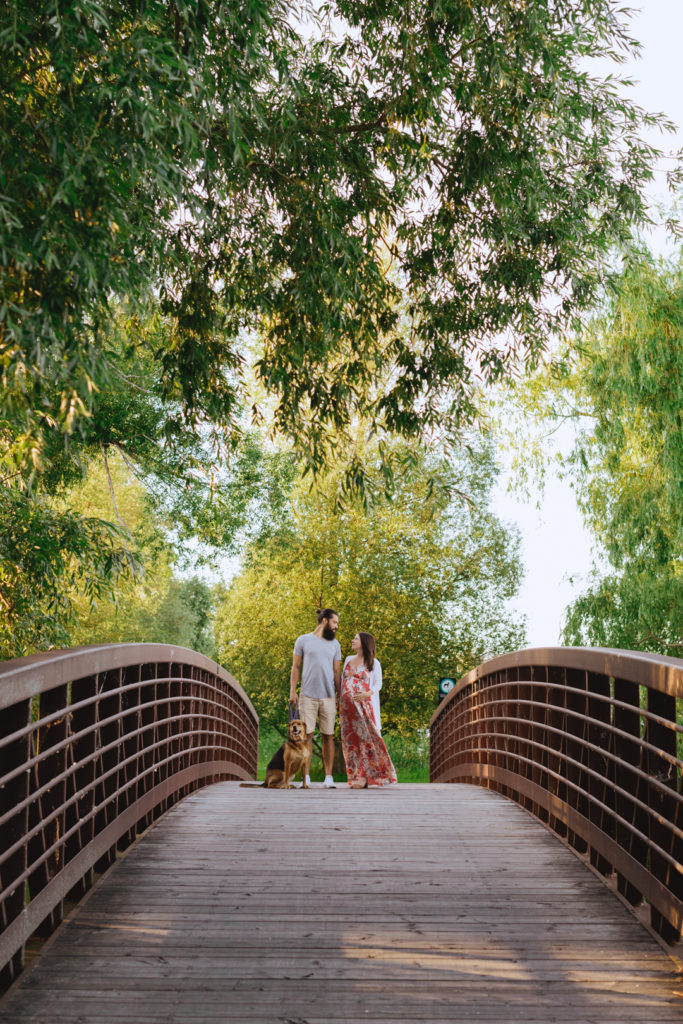 Expecting parents and their dog standing on bridge underneath the trees