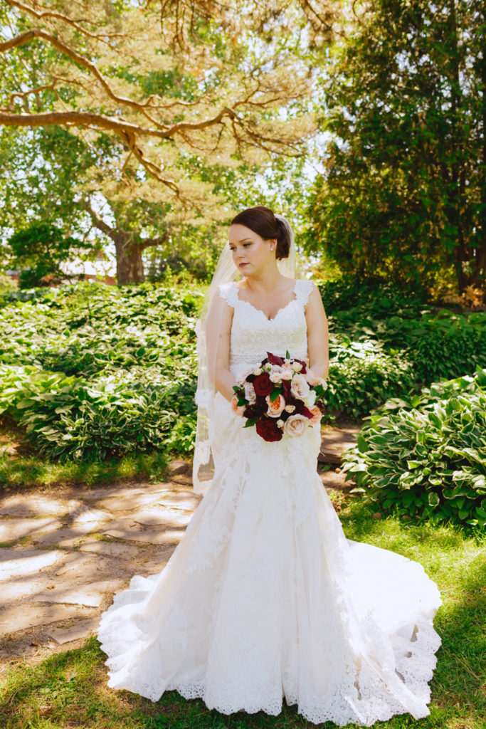 bride underneath green trees holding a red and blush bouquet