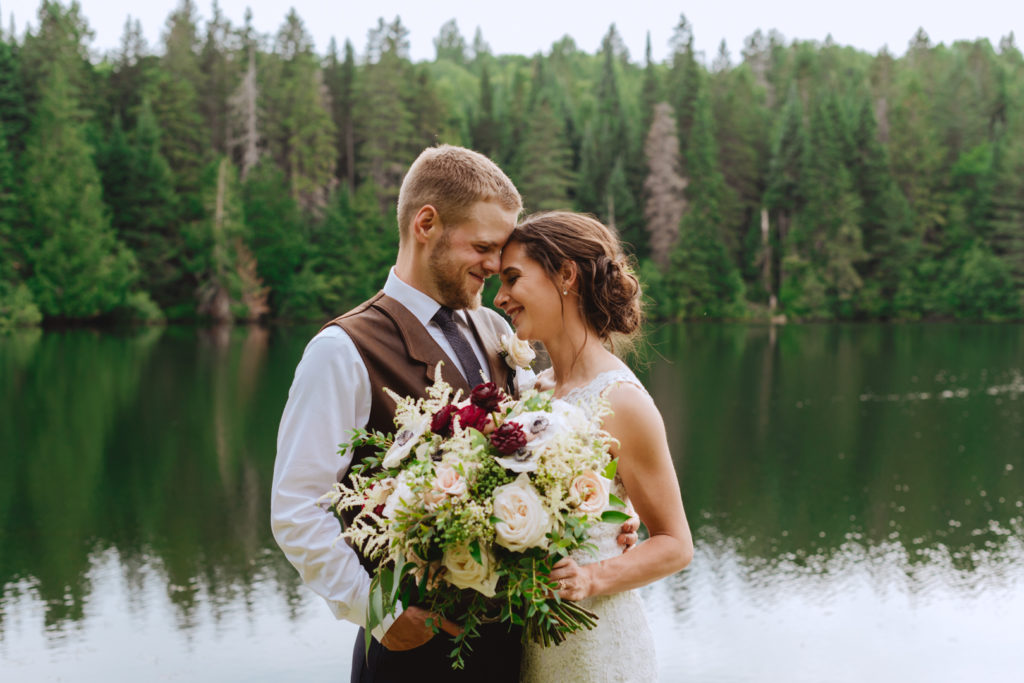 bride and groom cuddling by the water with evergreen trees in the background