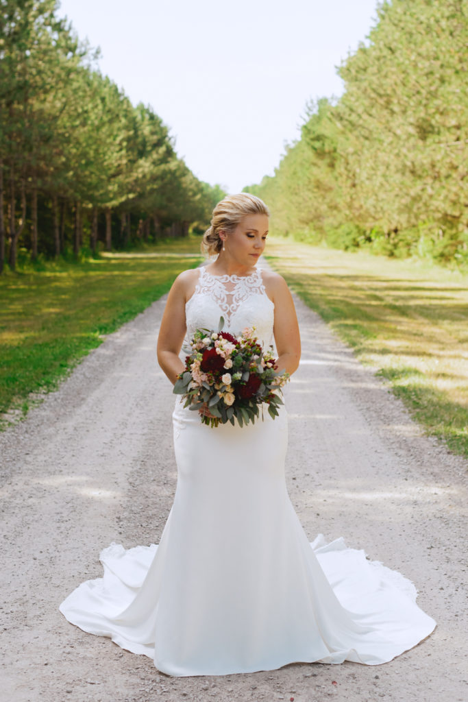 bride standing on dirt road surrounding by trees holding bouquet